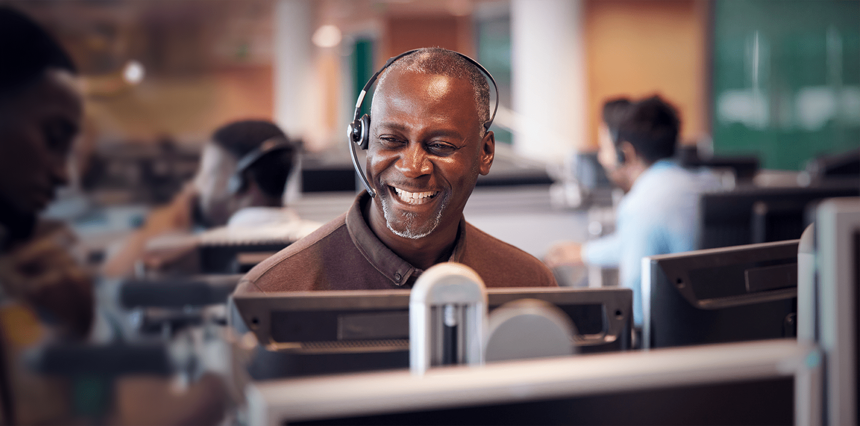 A salesperson sitting at a computer and wearing a headset is smiling while helping a customer find the automation part they need.